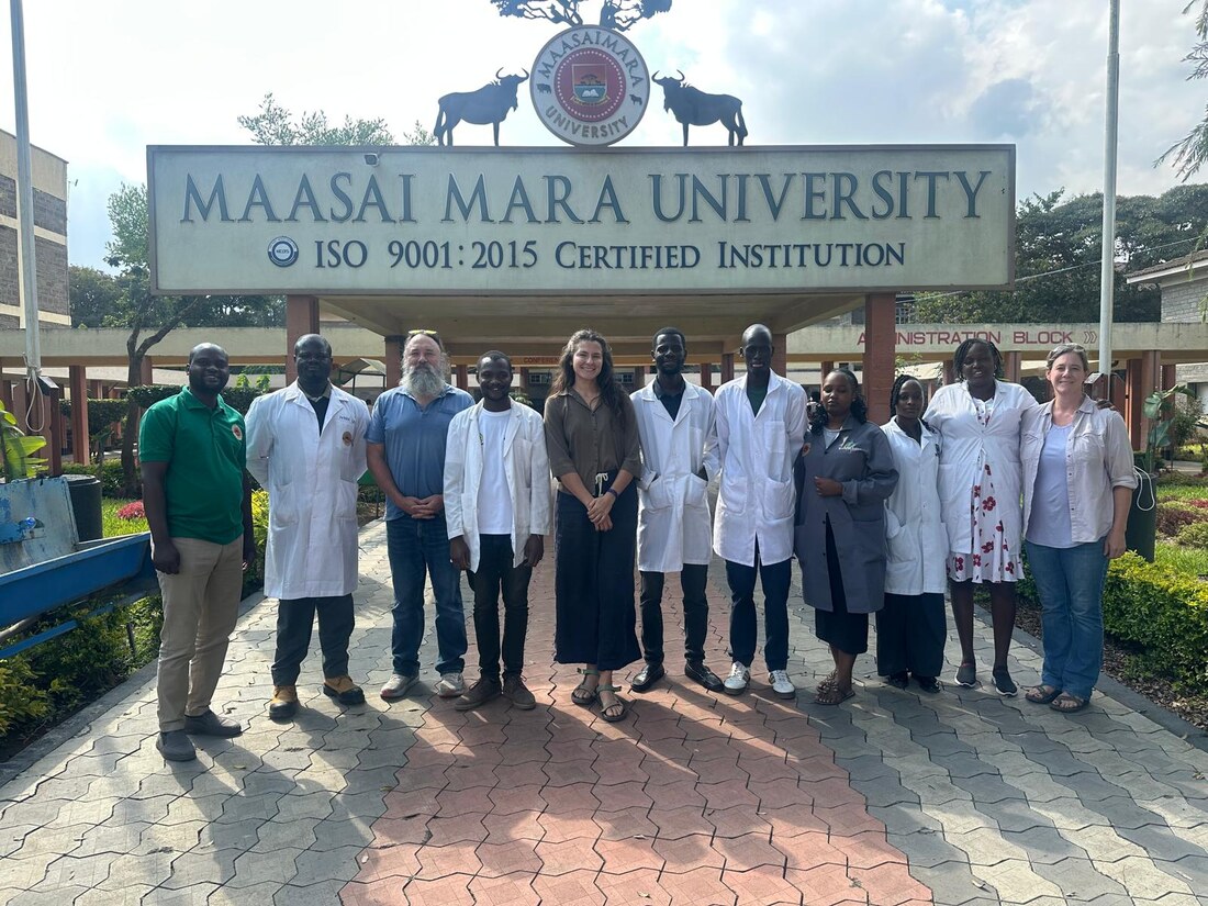 The team with Maasai Mara University staff and students in lab coats beneath the university entrance sign